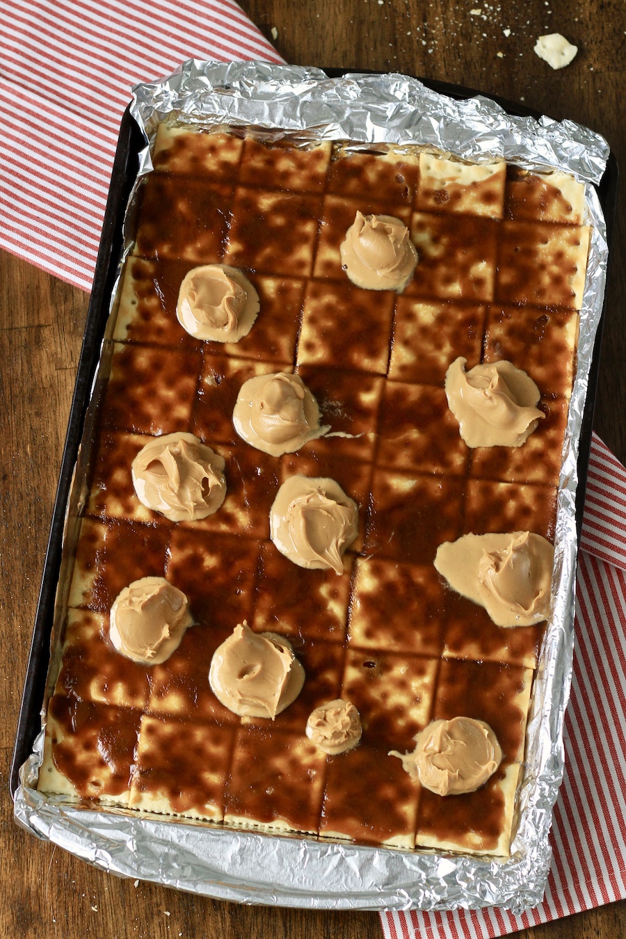 A baking sheet lined with foil topped with saltines covered with toffee topped with peanut butter.