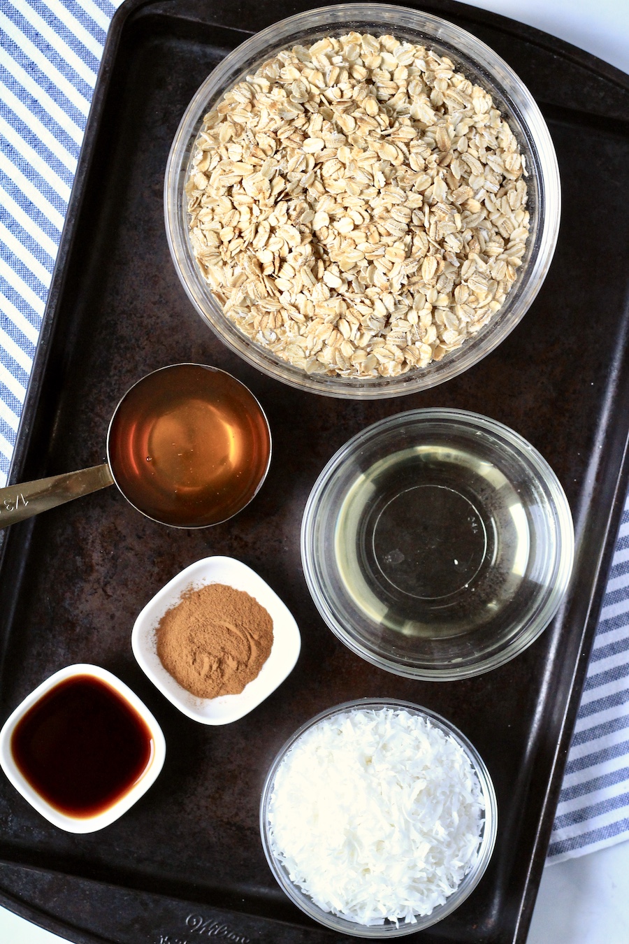 A dark, rimmed baking sheet with the ingredients in bowls for coconut granola.