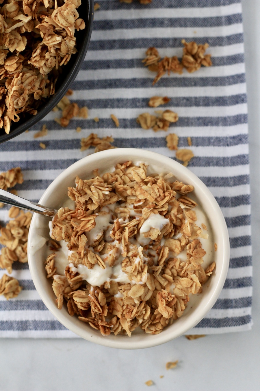 A top down photo of a white bowl with yogurt topped with crunchy homemade coconut granola with a silver spoon to the left.