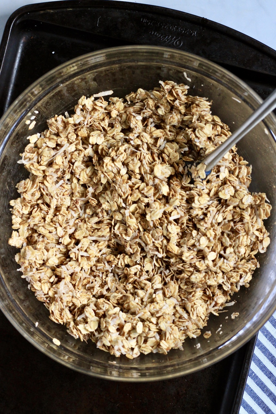 A glass bowl on a dark rimmed cookie sheet with the mixed ingredients for coconut granola and a silver spatula.