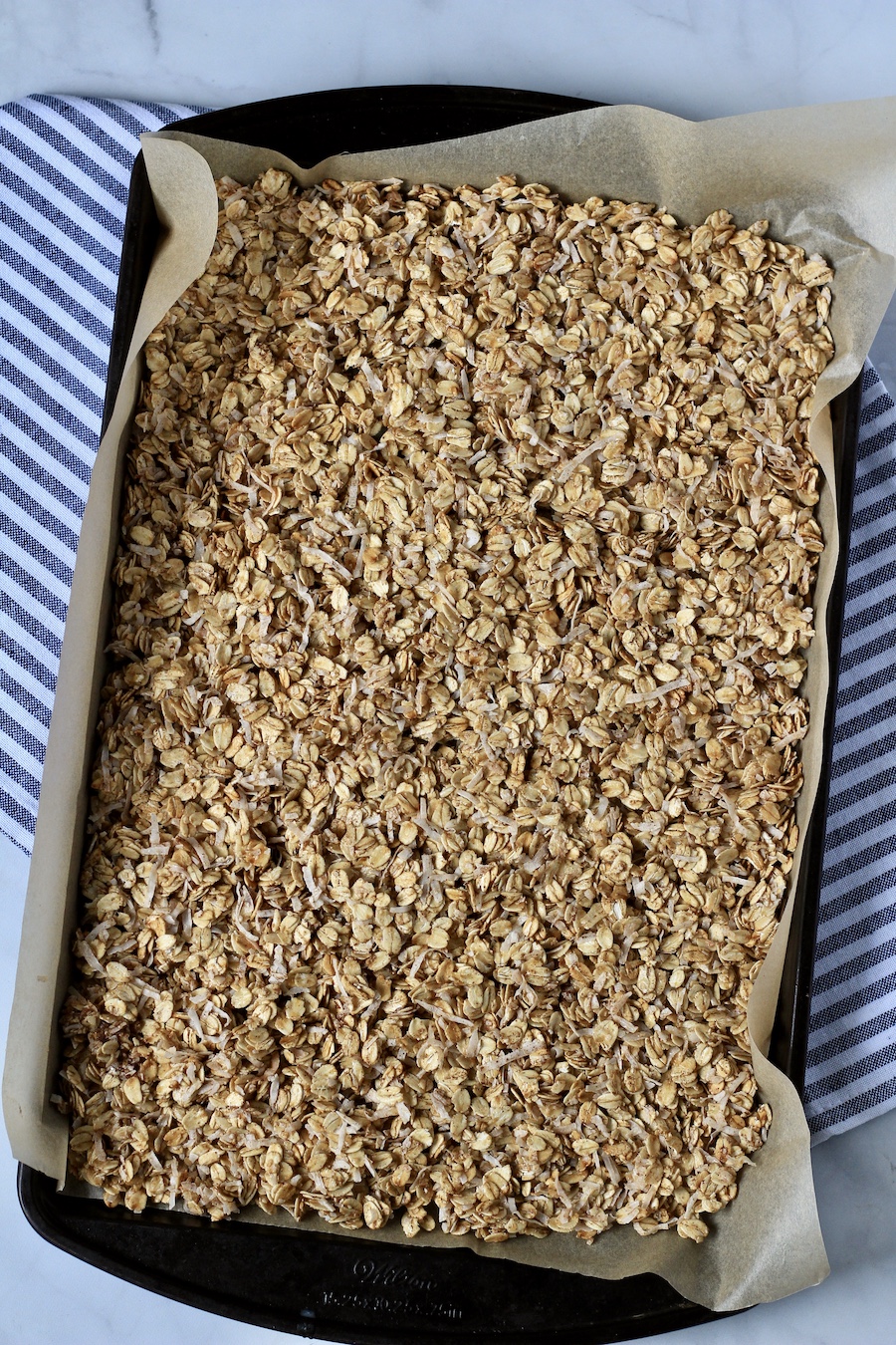 A parchment paper lined baking sheet with unbaked coconut granola on top of a white and blue striped towel.