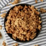 A top down picture of a blue bowl filled with coconut granola on a blue and white towel with pieces of granola around.