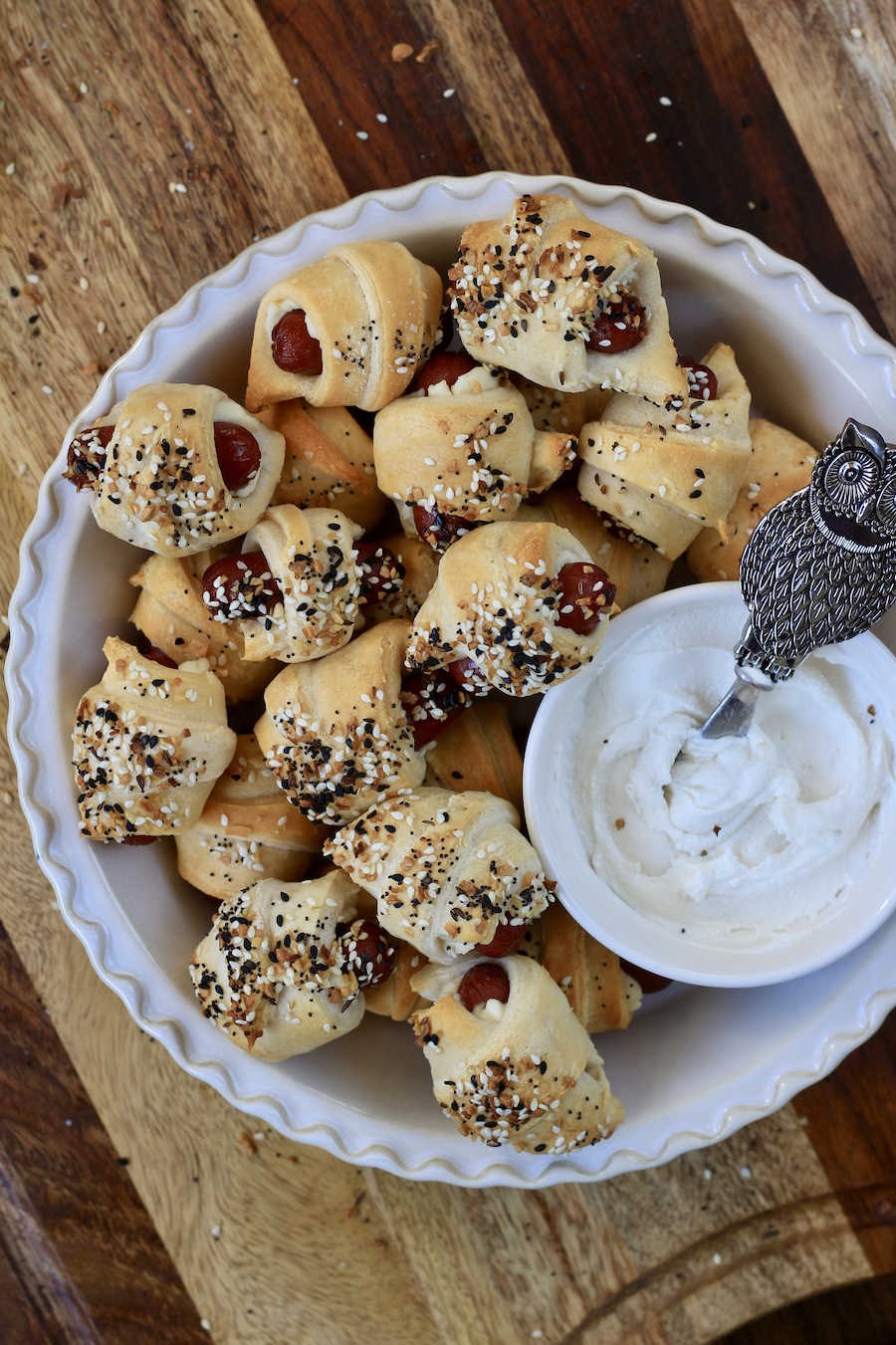 A pie pan full of baked everything bagel pigs in a blanket with a bowl of kite hill cream cheese.