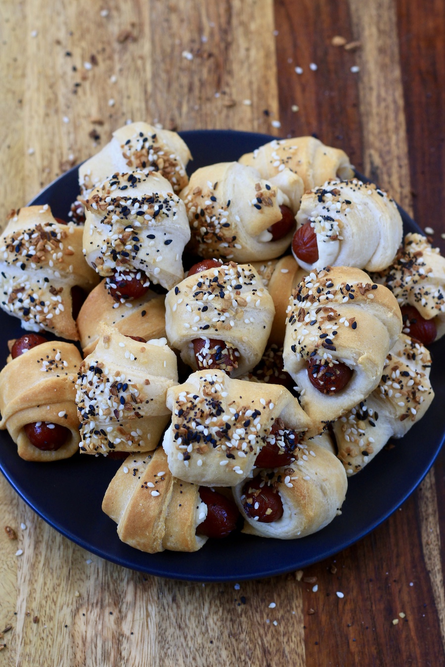 A small blue plate with a pile of everything bagel pigs in a blanket on a wooden cutting board.