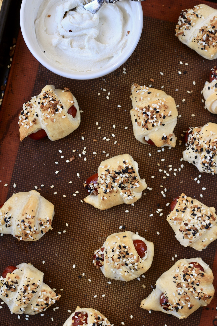 Everything bagel pigs in a blanket on a lined baking sheet with a bowl of kite hill cream cheese with a metal owl spreader.