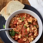 A top down photo of a white bowl filled with minestrone soup on a blue plate with a slice of bread.