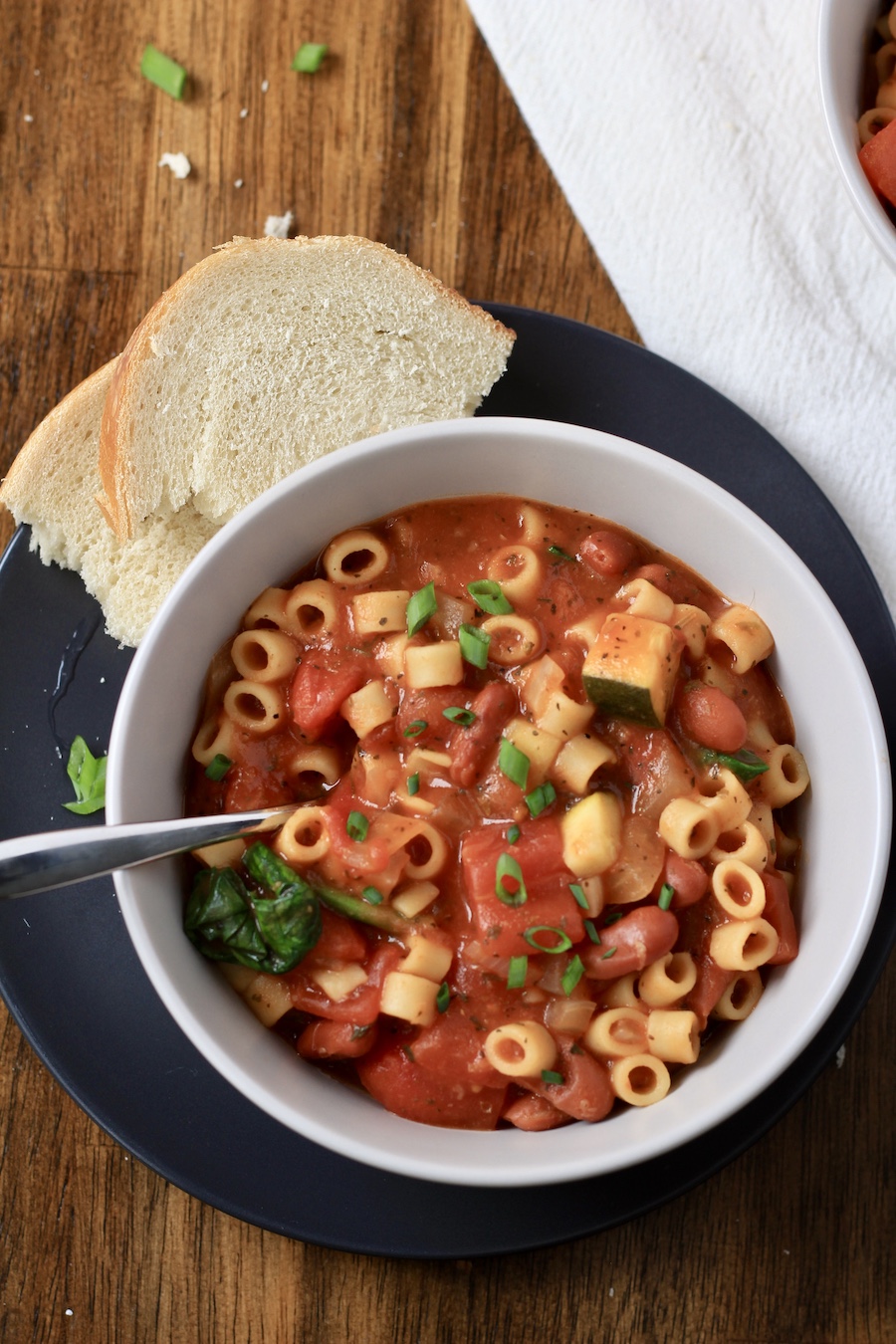 A top down photo of a white bowl filled with minestrone soup on a blue plate with a slice of bread.