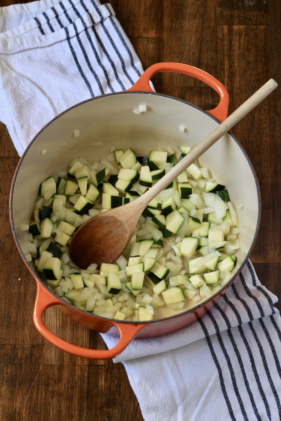 Zucchini, onion, and garlic cooking in an orange dutch oven with a wooden spoon on a white and blue dish towel.