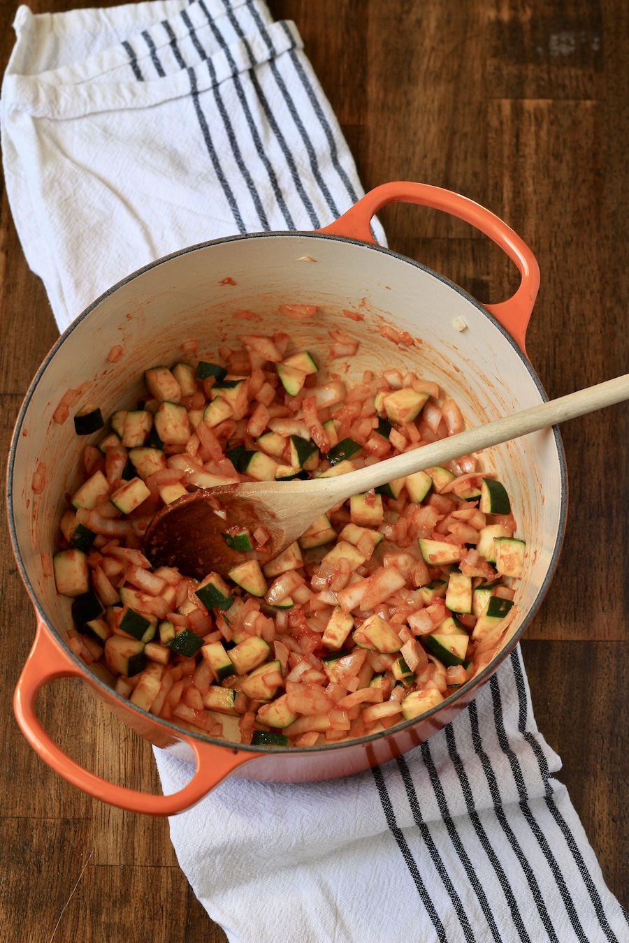 Cooked zucchini, onions, and garlic with tomato paste in an orange dutch oven pot on a white and blue striped towel.
