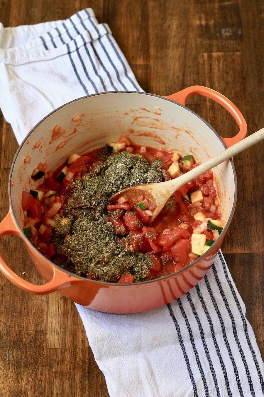 Vegetables topped with spices in an orange dutch oven with a wooden spoon on a white and blue striped towel.