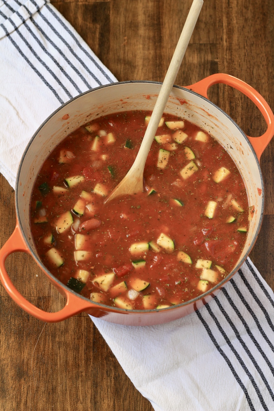 Minestrone soup after adding pasta in an orange dutch oven with a wooden spoon on a white and blue dish towel.