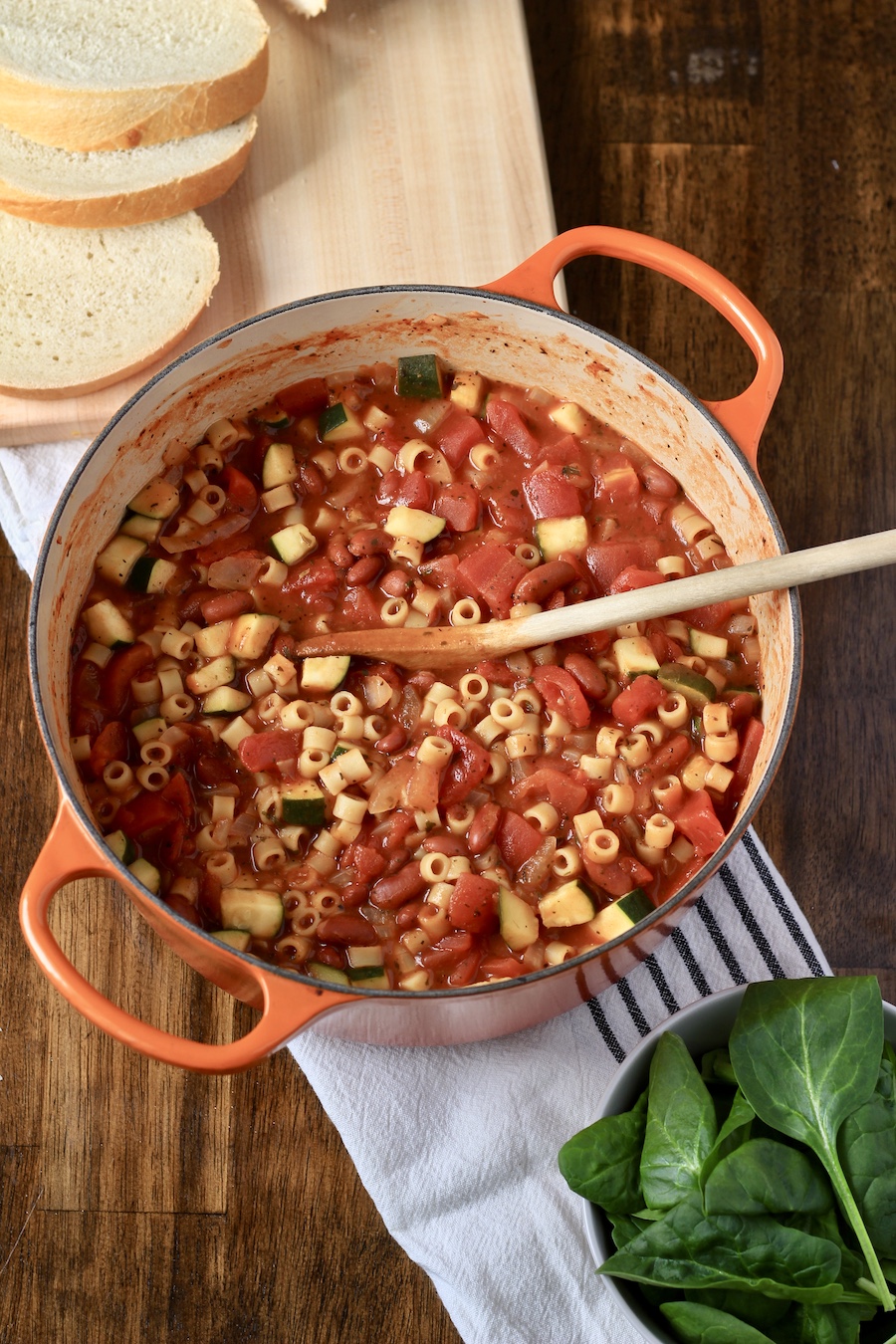 An orange dutch oven with a wooden spoon on a white and blue dish towel with a bowl of spinach to the right and a cutting board with bread to the top left.