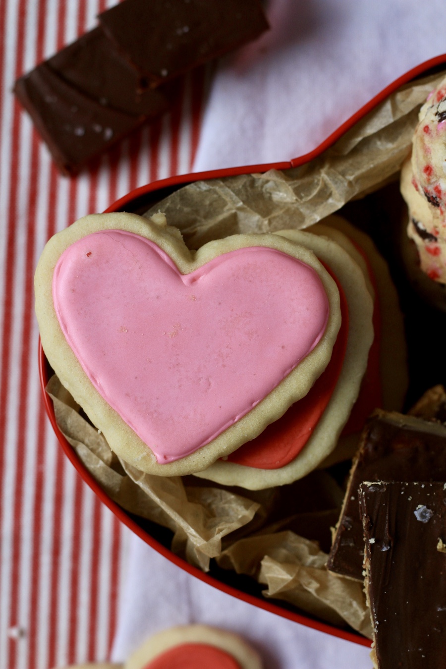 A pink iced sugar cookie in the left of the dessert box lined with parchment paper on a white towel.