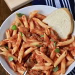 A white bowl with penne pasta mixed with Arrabbiata meat sauce with a slice of bread on the right side.