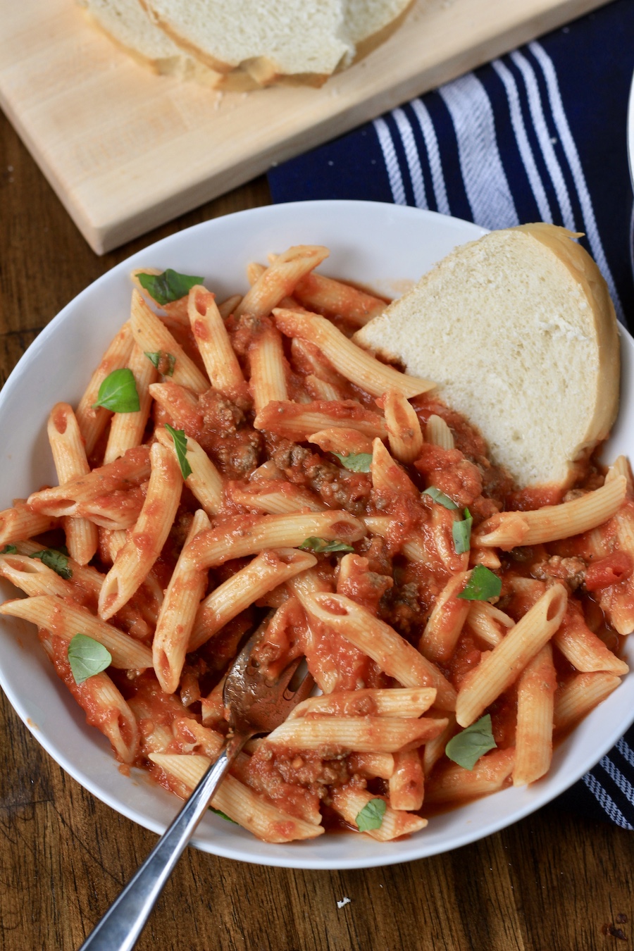 A white bowl with penne pasta mixed with Arrabbiata meat sauce with a slice of bread on the right side.