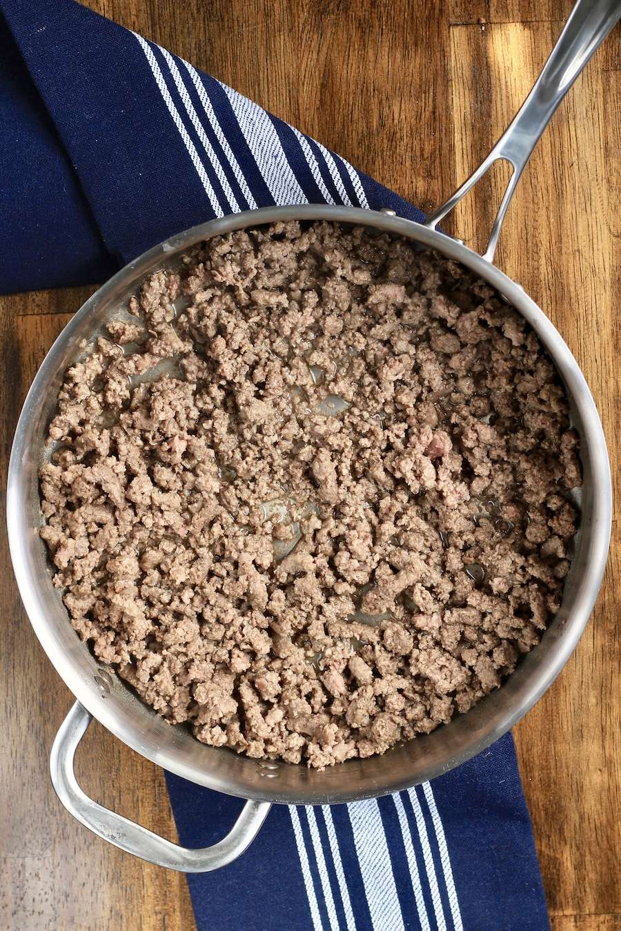 A skillet with browned ground beef on a wooden counter with a blue towel.
