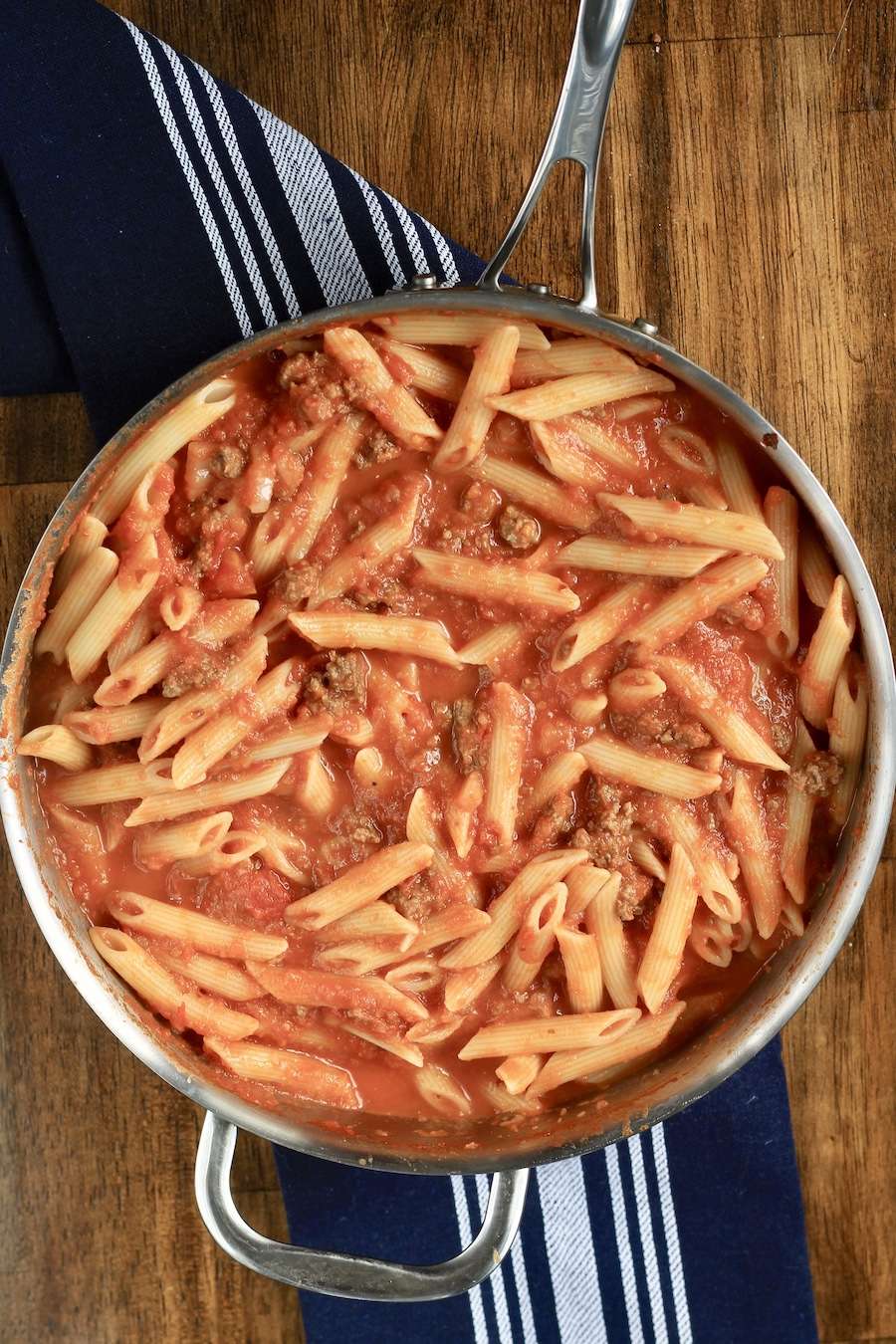 Penne pasta mixed in Arrabbiata meat sauce in a skillet on a blue towel on a wooden counter.