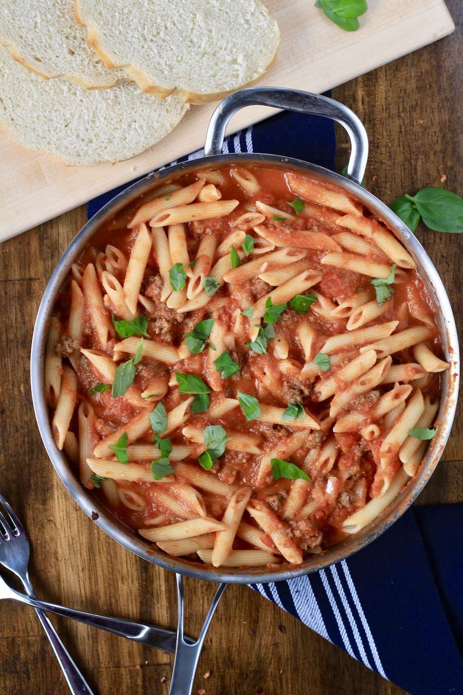 A skillet with Arrabbiata pasta and a cutting board in the back with slices of bread in the back.