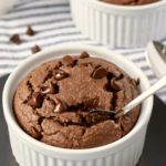 White ramekins filled with chocolate baked oats topped with chocolate chips and a silver spoon in the baked oats on a white and blue striped towel.