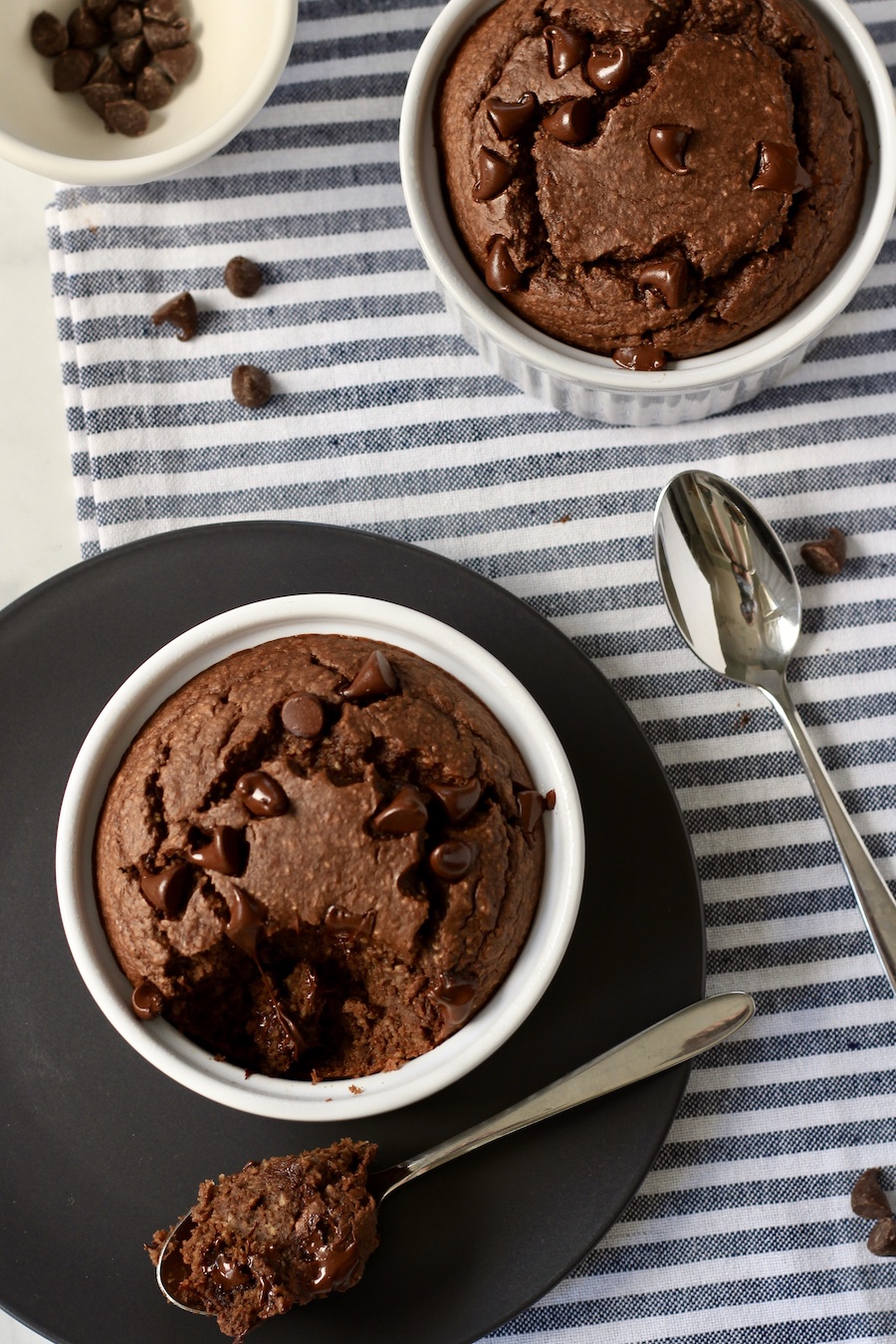 A blue plate with a white ramekin and a spoon with a chunk of chocolate baked oats taken out with a white and blue striped towel.