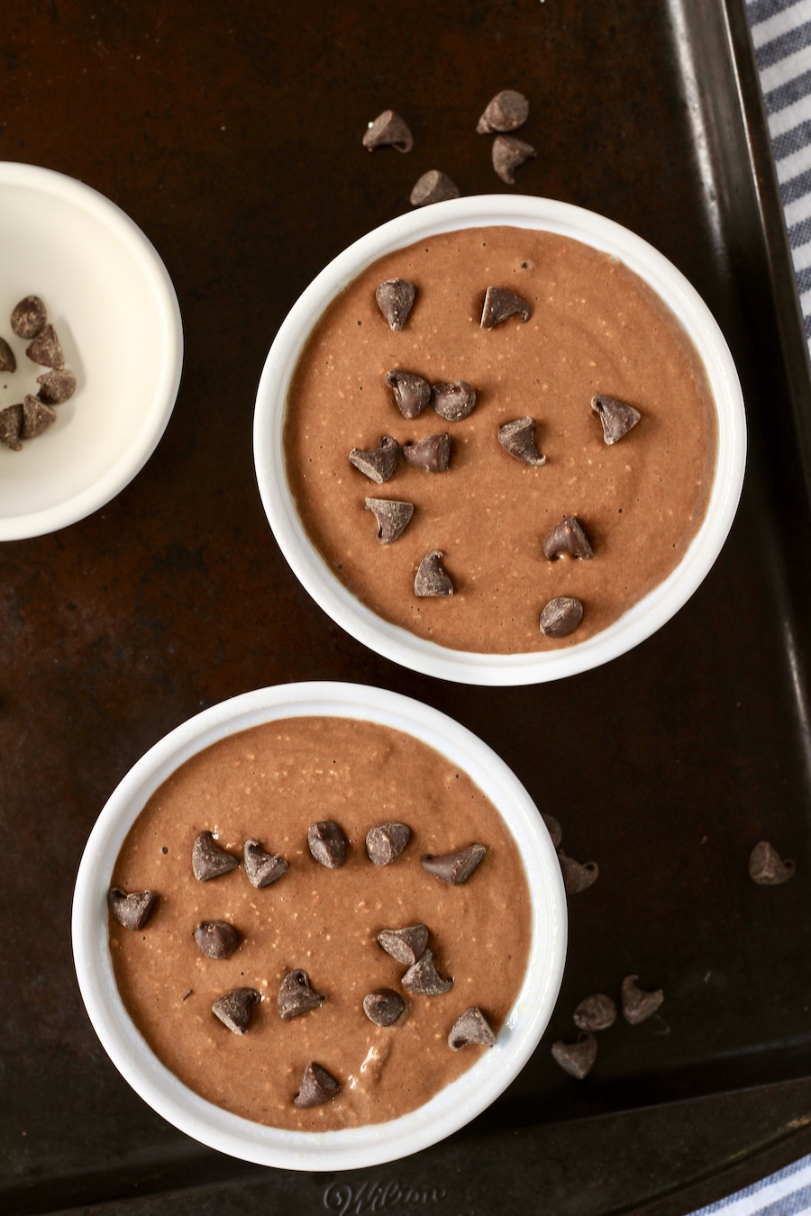 Two white ramekins on a baking sheet with chocolate baked oats before baking.