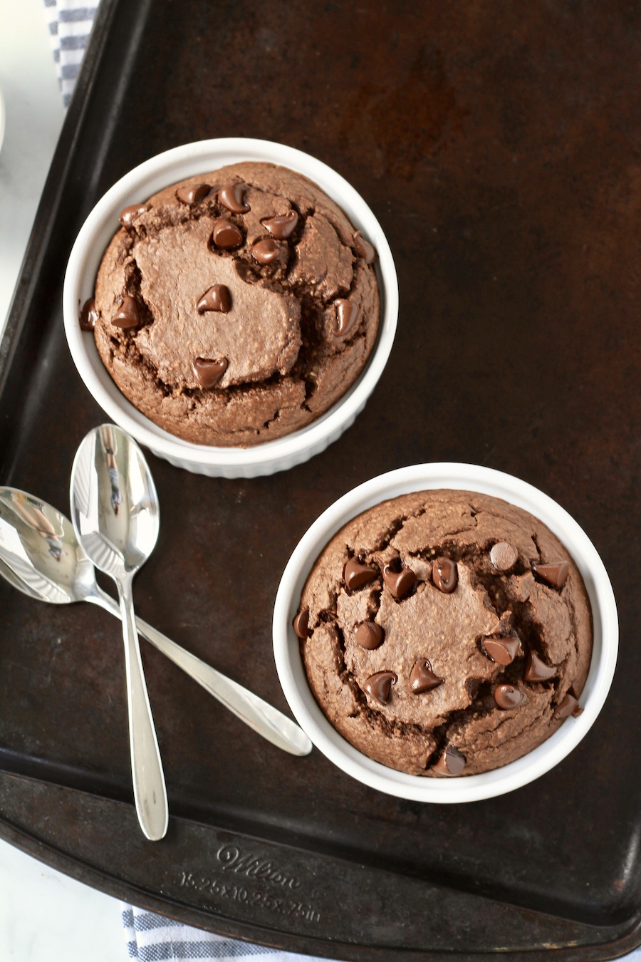 Top down photo of two ramekins with chocolate baked oats on a baking sheet with two spoons to the left.