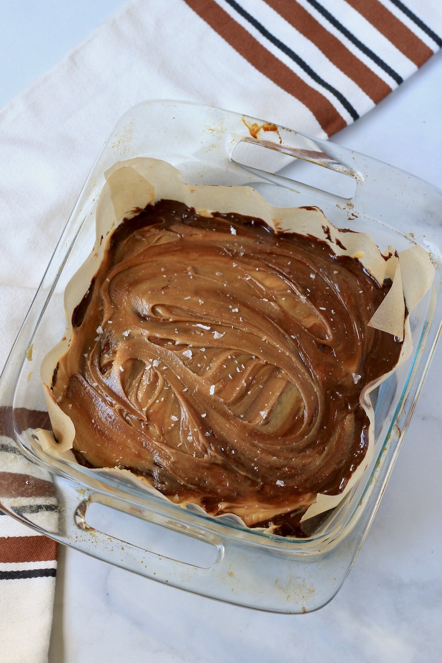 A square baking dish lined with parchment paper and filled with chocolate peanut butter swirled fudge.