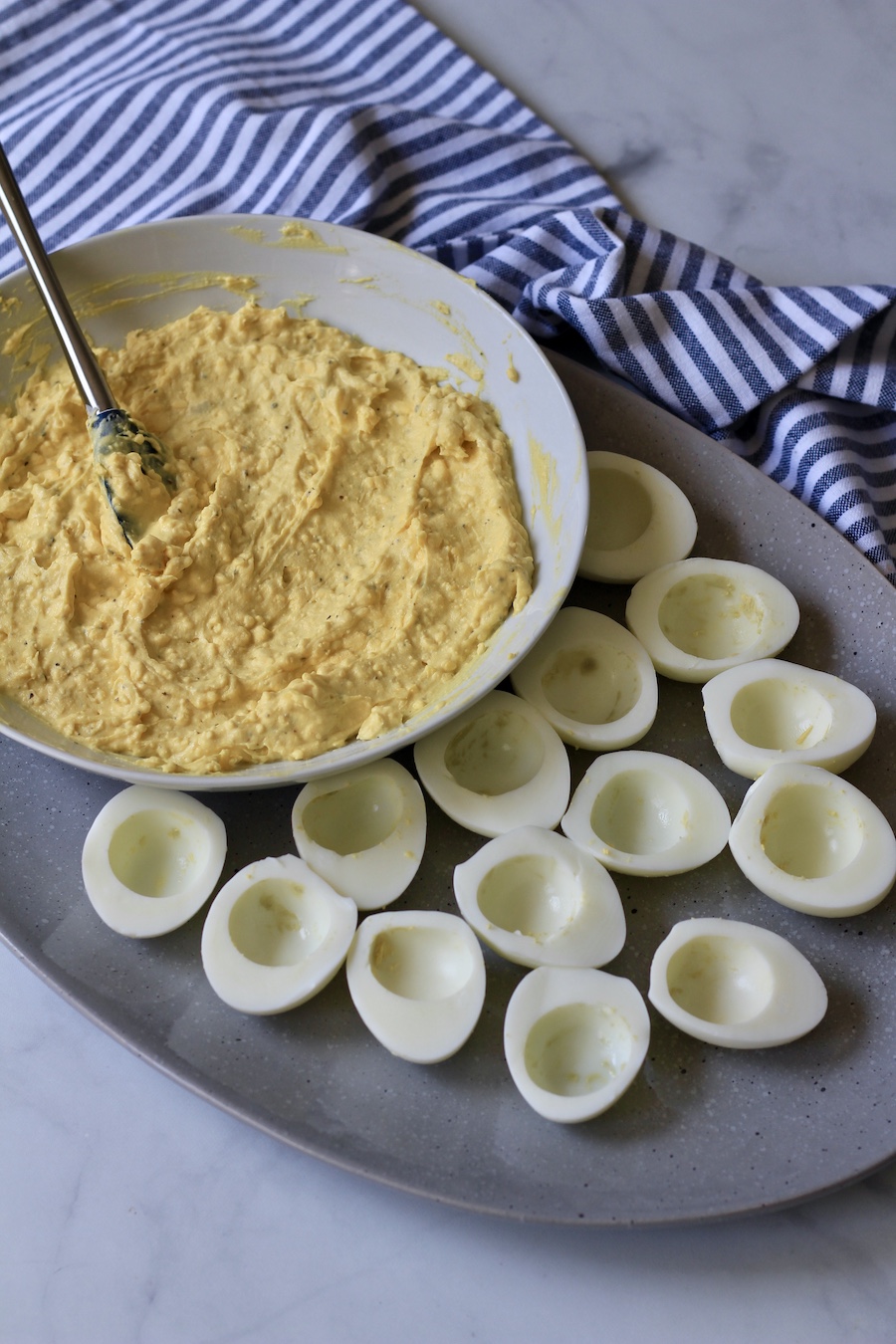 Deviled egg filling in a white bowl with a spoon and hard boiled egg white halves on a platter with a blue and white striped towel.
