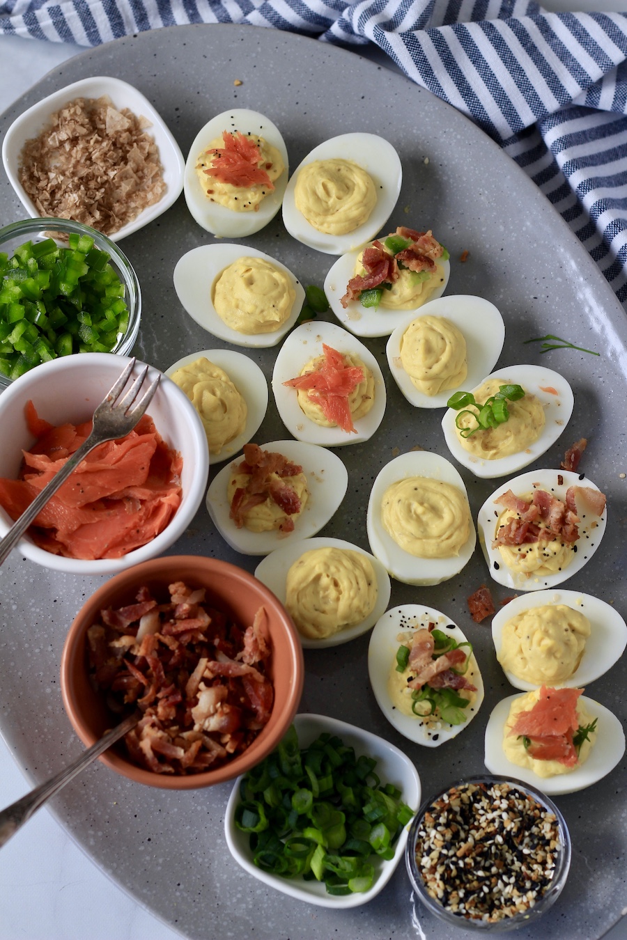 A deviled egg board with deviled eggs topped with different toppings and bowls of toppings on the left side of the platter.