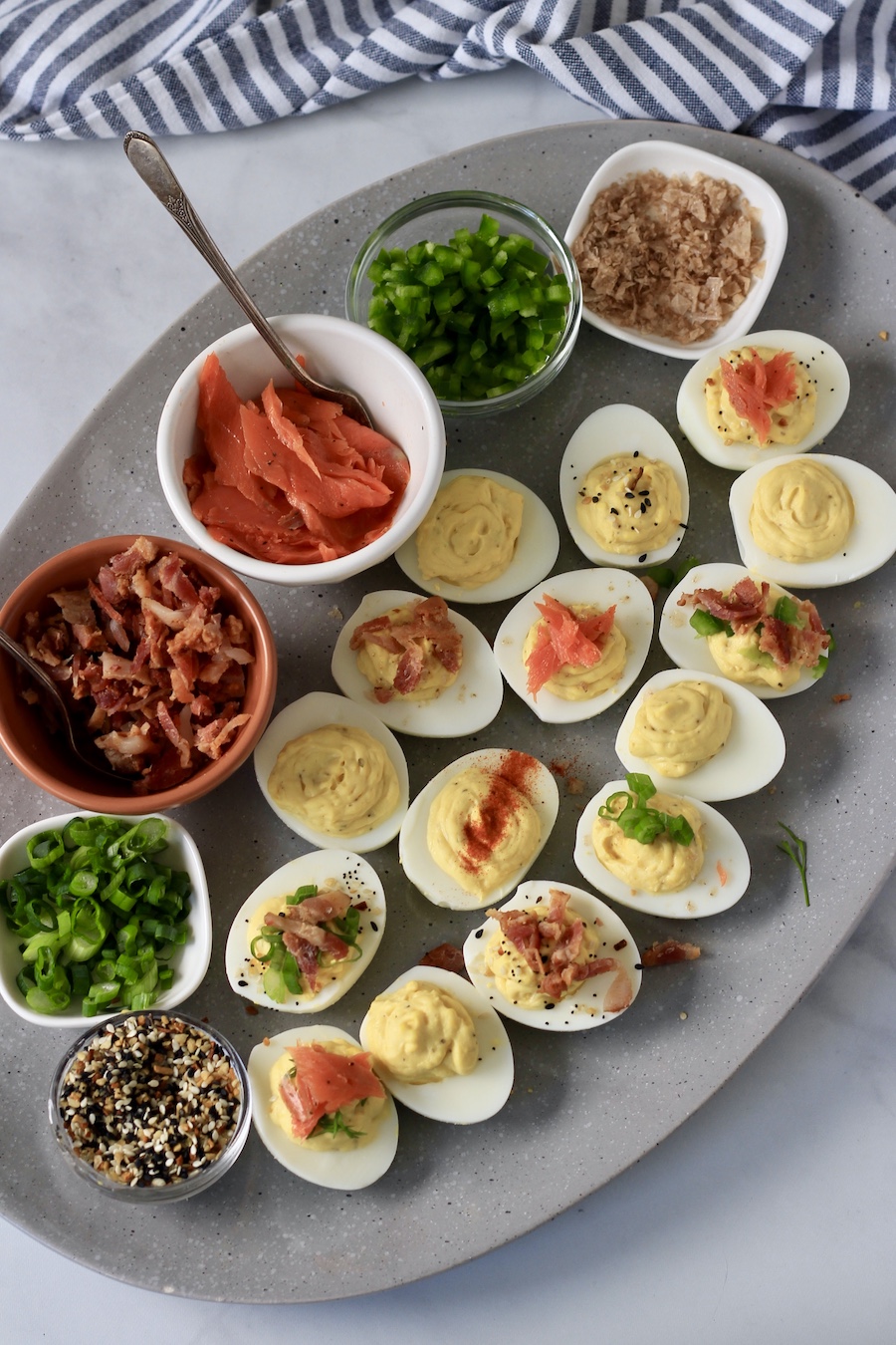 A grey platter from the top down with deviled eggs in the front and bowls of toppings in the back with a blue and white towel in the back right.