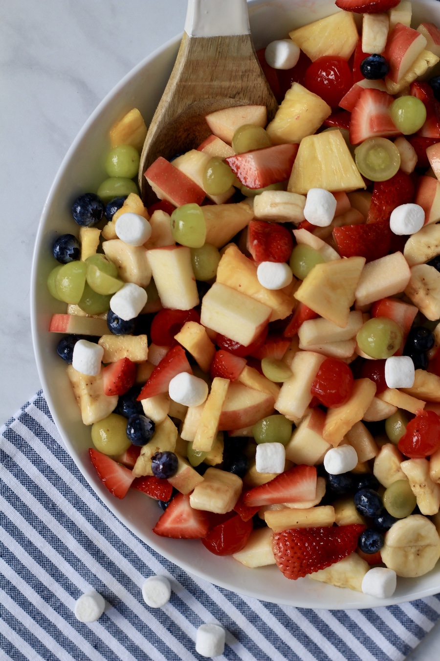 The left side of a white bowl of fruit salad with a brown spoon and some marshmallows on the counter.