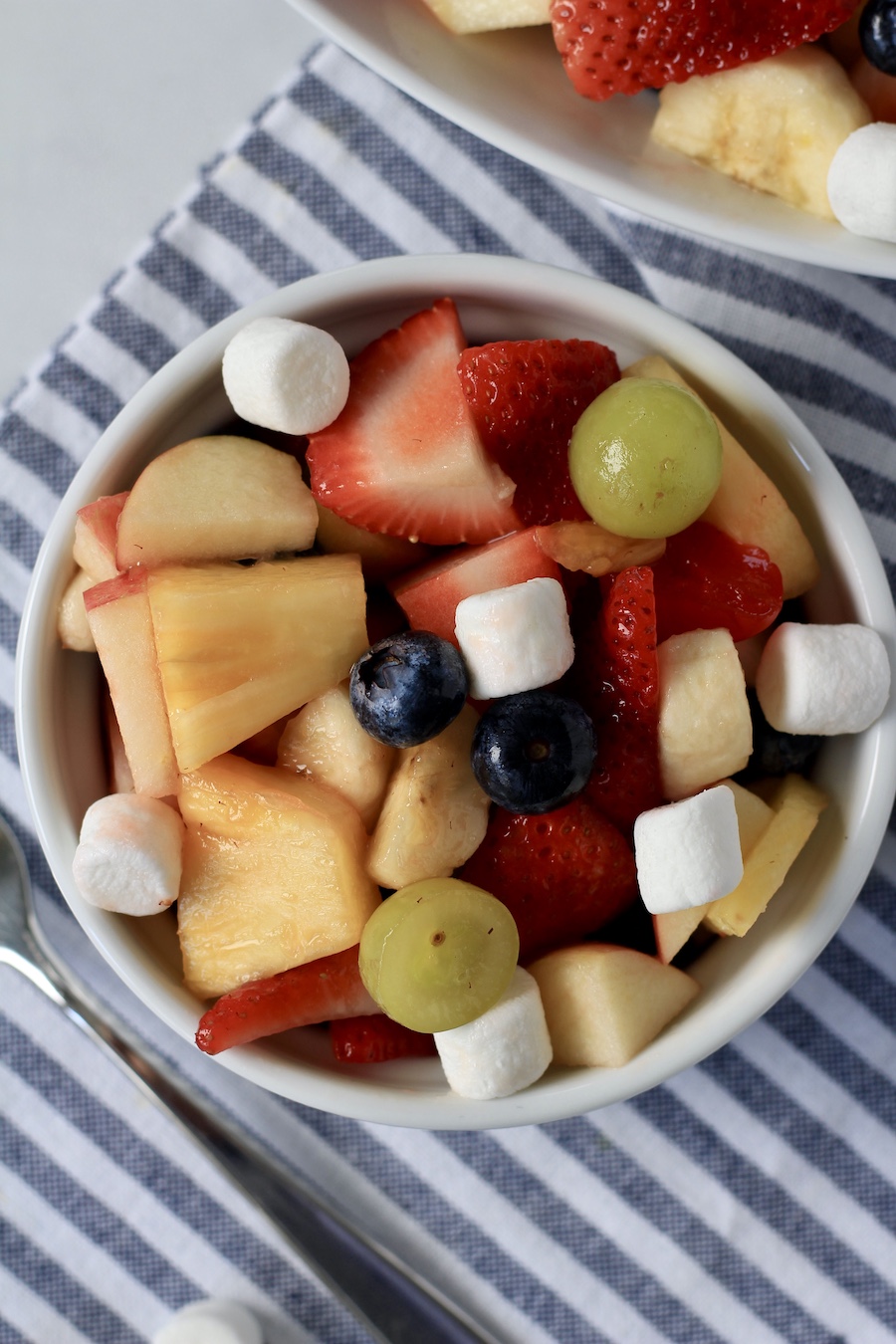 A white ramekin with fruit salad topped with marshmallows on a white and blue towel.
