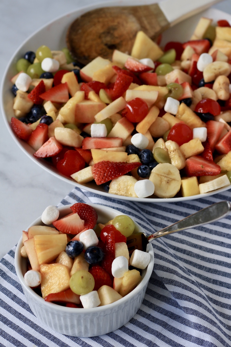 A small white bowl with fruit salad and a large bowl of fruit salad in the back with a wooden spoon.