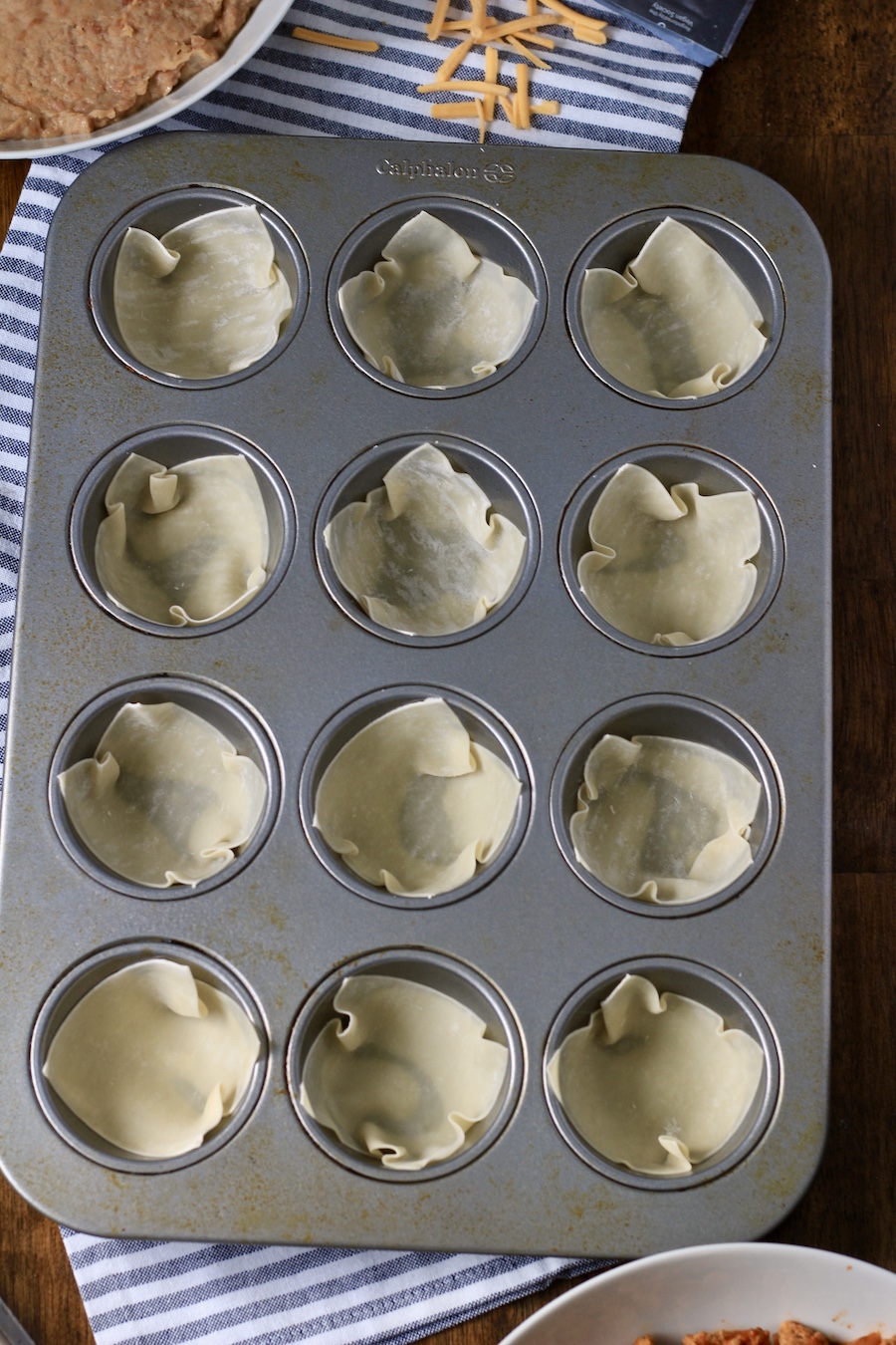 A muffin tin with wonton wrappers on a white and blue towel.