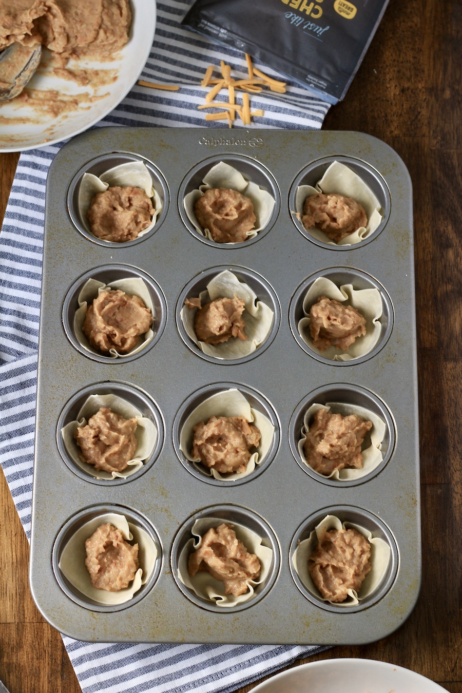 Wonton wrappers topped with refried beans in a muffin tin on a white and blue towel.