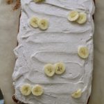 A full banana slab cake with cinnamon frosting and banana slices on top on a piece of parchment paper.