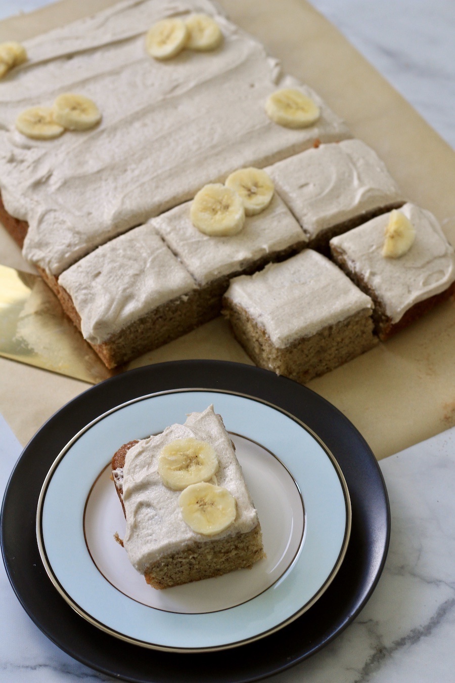 A corner of banana slab cake on a blue and white plate with the full slab cake in the back.