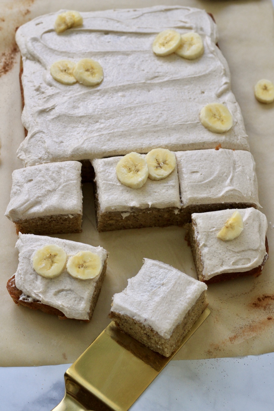 Parchment paper with a banana snack cake on top and 6 slices cut with a  gold spatula under a slice.