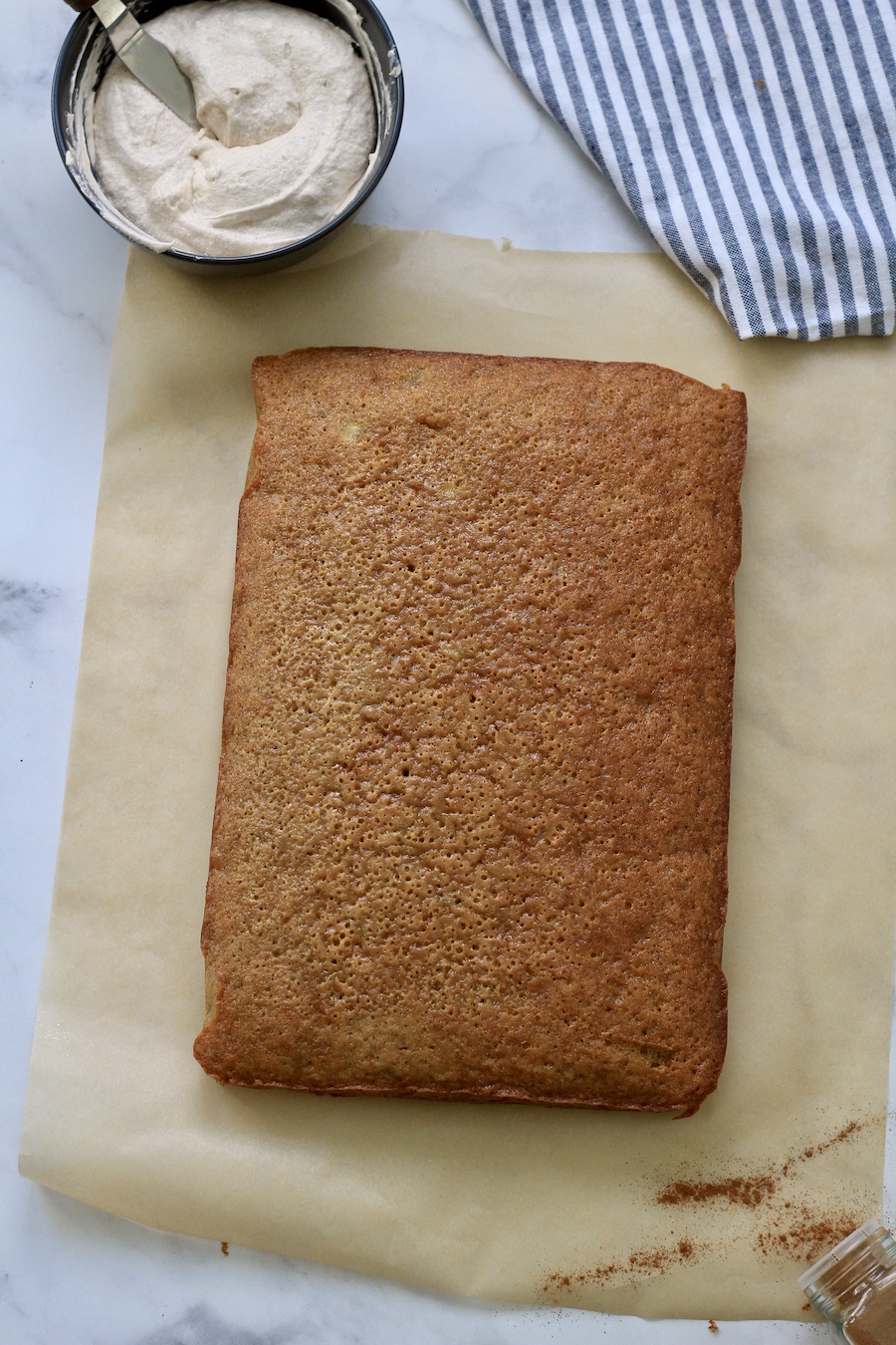 A banana slab cake on parchment paper with a bowl of frosting in the top left.