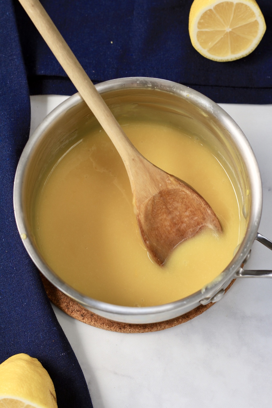 A sauce pan with lemon curd and a wooden spoon and a lemon in the bottom left corner and another lemon in the top right corner.