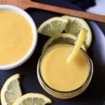 A blue plate with two bowls of lemon curd with slices of lemons and a wooden spatula in the back.