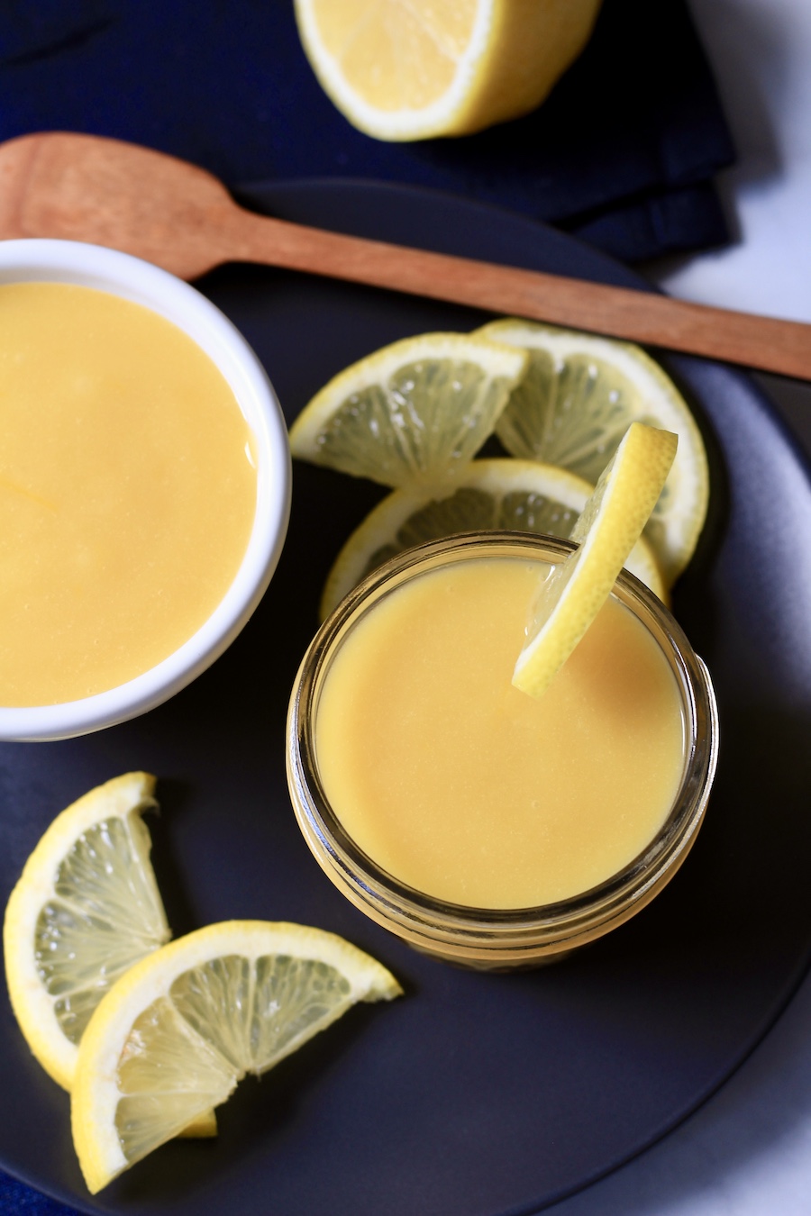 A blue plate with two bowls of lemon curd with slices of lemons and a wooden spatula in the back.