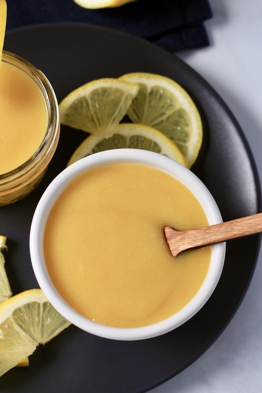A white bowl with lemon curd and a small wooden spatula in the bowl with lemon wedges.