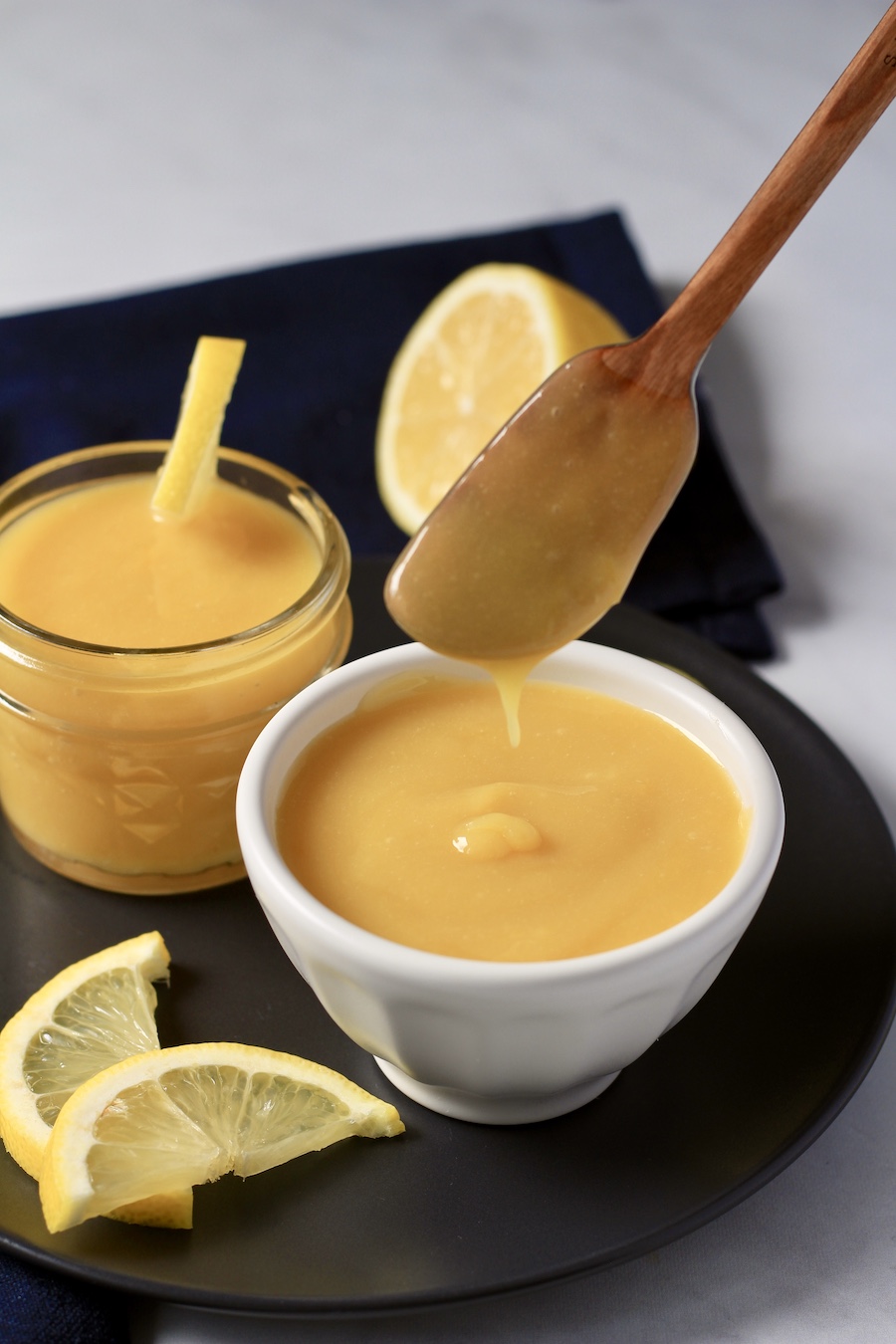 A white bowl of lemon curd with a small wooden spatula dipped in and small lemon wedges in the front.