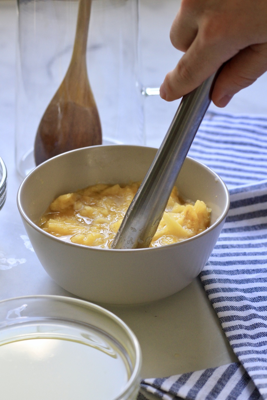 A hand holding a muddler smashing pineapple in a bowl on a white and blue towel.