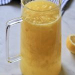 A glass pitcher of pineapple lemonade on a counter with a white and blue striped towel in the back.