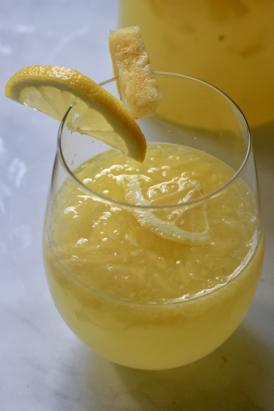 A glass of pineapple lemonade with a lemon wedge and a small piece of pineapple on the rim.