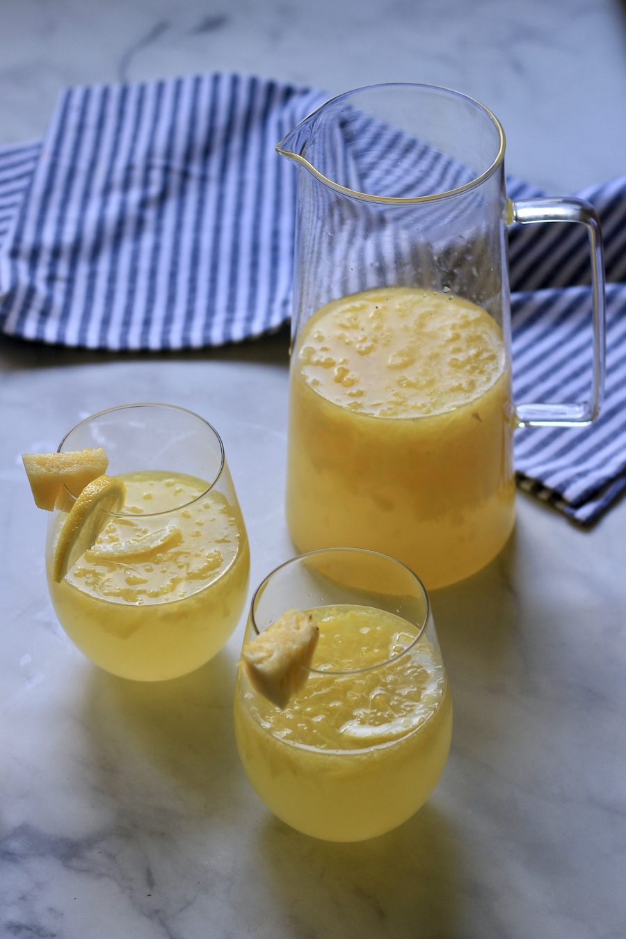 A glass pitcher of lemonade in front of a white and blue striped towel with two glasses of pineapple lemonade in the front.
