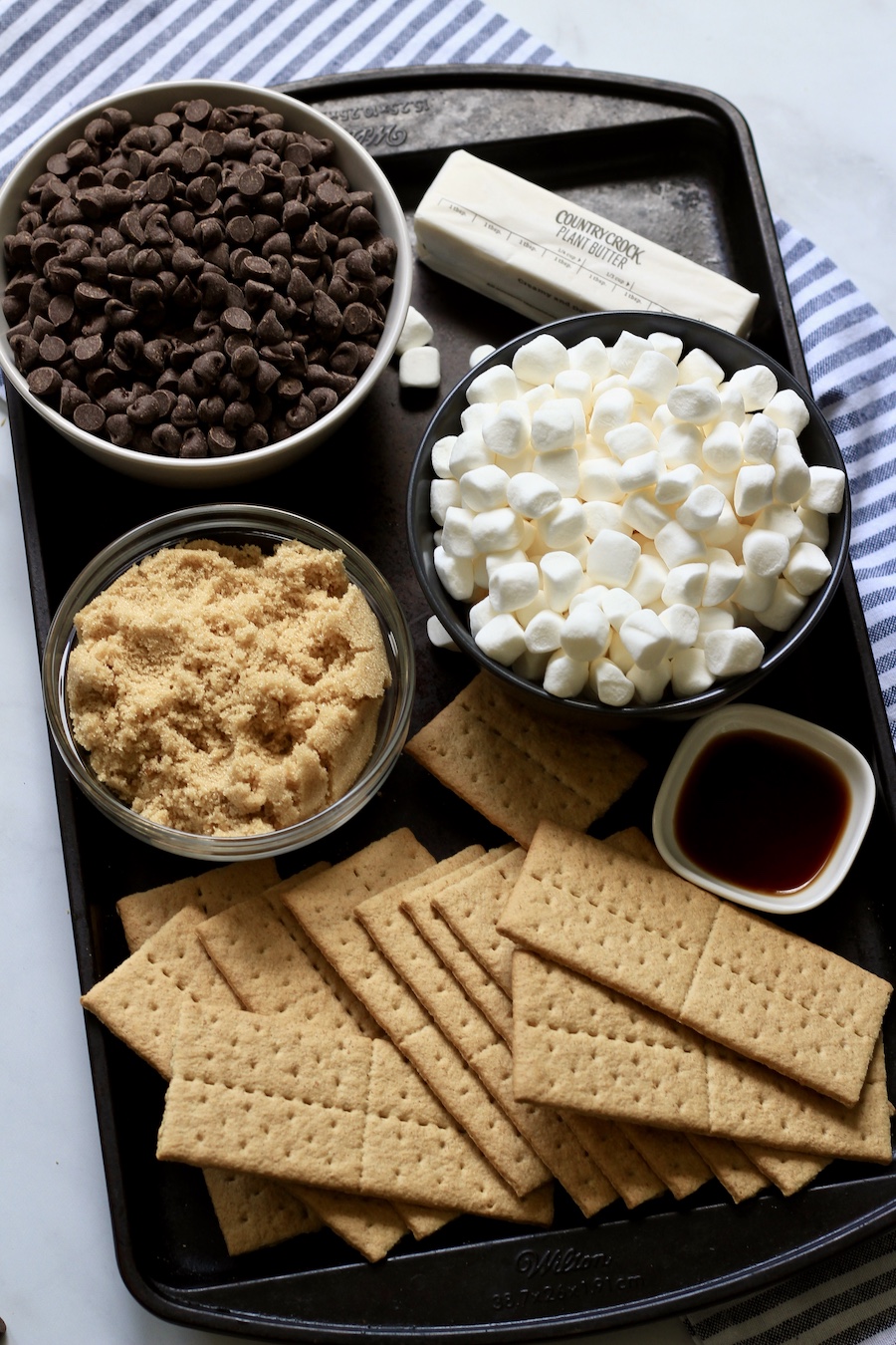 A rimmed baking sheet with ingredients for smores toffee with a white and blue dish towel in the back.