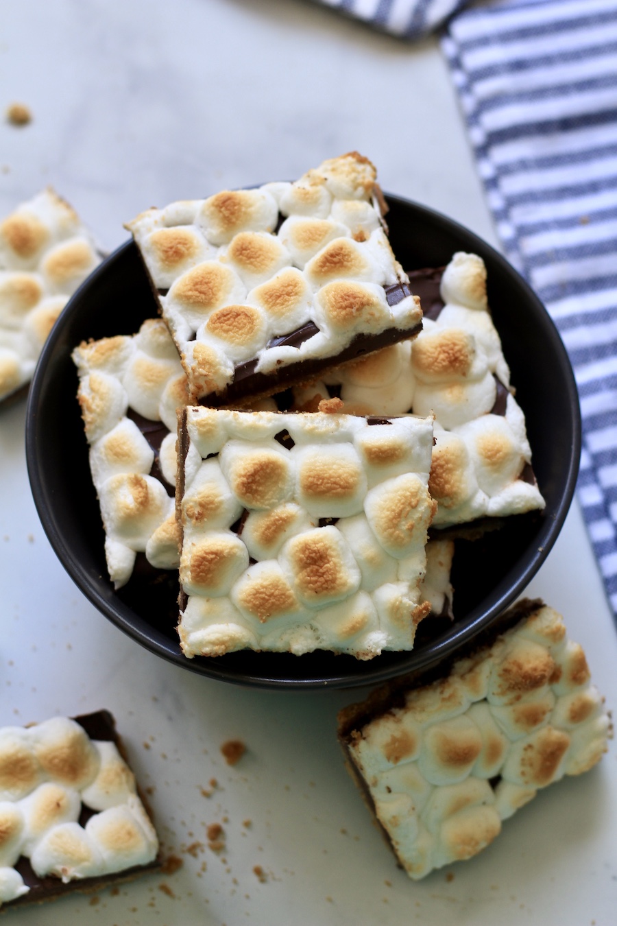 A blue bowl filled with pieces of s'mores toffee on a white counter with pieces of s'mores toffee around it.